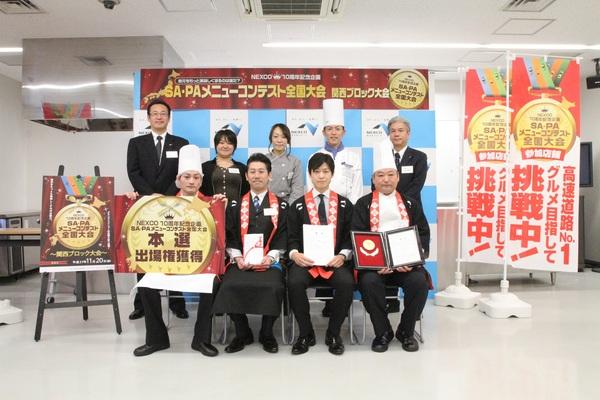 サービスエリア・パーキングエリアメニューコンテスト全国大会関西ブロック大会の看板の前で、グランプリを受賞した「舞鶴若狭自動車道 西紀サービスエリア上り線アイランドフューチャーコーポレーション株式会社」の皆さんが、本選出場権獲得とグランプリの盾、賞状、賞金を持って写っている記念写真