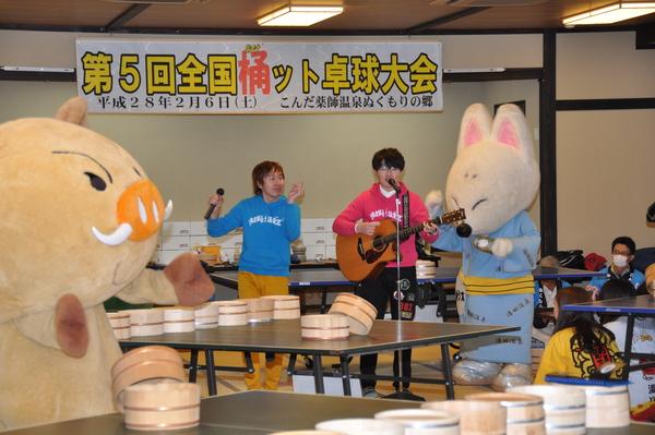 まるいのときつねの着ぐるみの間でギターとマイクを持った男性二人が歌っている写真