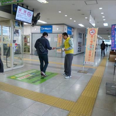 駅内で黄色の服を着た男性が黒色の服を着た男性に喫煙禁止のティッシュを配っている写真