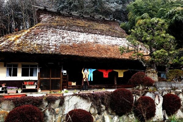かやぶき屋根に薄っすら雪がかかっている軒下にエプロンを着た女性がカラフルな色の服を干している最優秀賞受賞の写真