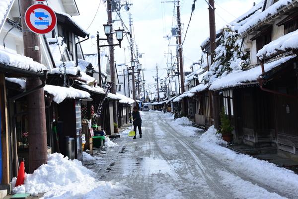 雪が積もった妻入商家群沿いの道路を黄色いスコップで雪かきをしている優秀賞の写真