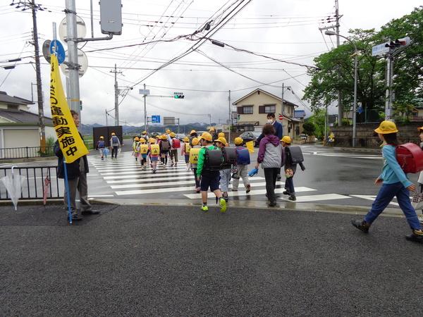 保護者が黄色のあいさつ運動実地中の旗を持ち、児童が横断歩道を渡っている写真