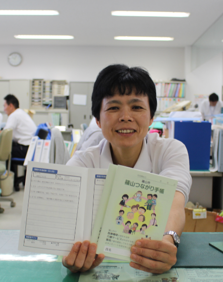 篠山つながり手帳を1冊と中身を開いた手帳を1冊両手に持っている女性職員の写真