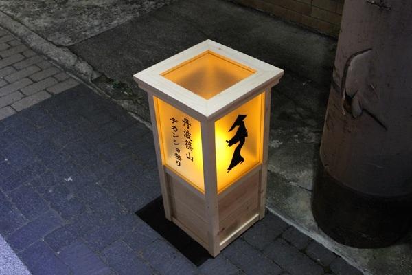 四角の木の板で天井をつくりその下に丹波篠山デカンショ祭りと黒字でかかれた隣に踊り姿のイラストがえがかれたあんどんに灯りがともされて道路に飾られている写真