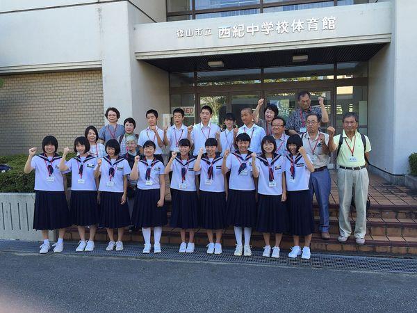 西紀中学校体育館の前で、ジュニアボランティアの生徒と関係者が2列に並び片手をガッツポーズをして写っている集合写真