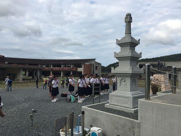 3重の塔のような石で作られている慰霊碑の前に、制服を着たジュニアボランティア生徒達が集まっている様子の写真