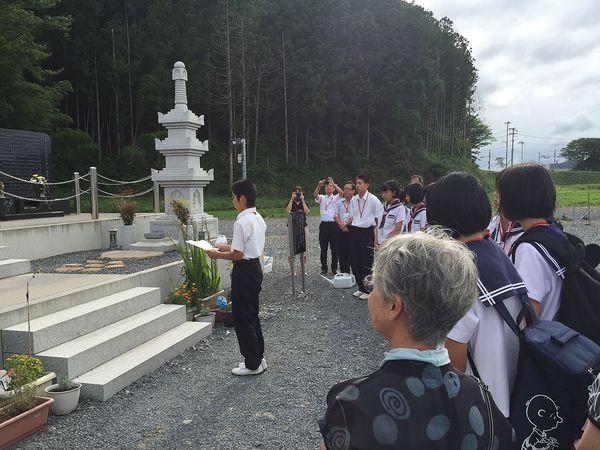 慰霊碑の前にジュニアボランティアの生徒と関係者が集まり、代表の男子生徒が追悼メッセージを読んでいる様子の写真