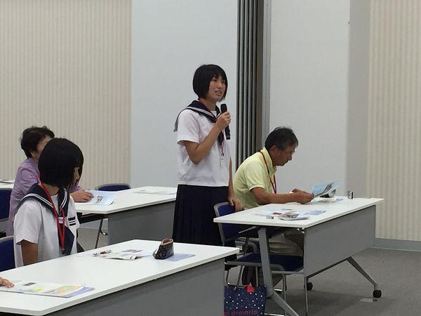 会議室で、女子生徒が自分の席から立ちマイクを持って発表をしている様子の写真
