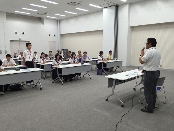 会議室にてジュニアボランティアに参加した生徒達は席に着いており、その中の男子生徒が席の横に立って、マイクを持った男性が話をしているのを聞いている様子の写真