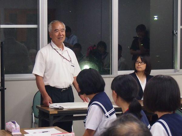 白いポロシャツを着た白髪の男性が立ち上がって話をしており、制服を着た女子生徒が話を聞いている後ろ姿の写真