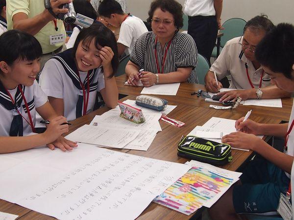 机の上に模造紙が置かれて女子生徒が文字を書いており、同じ机に生徒と年配の男性と女性も座っており、グループワークで話し合いをしている様子の写真