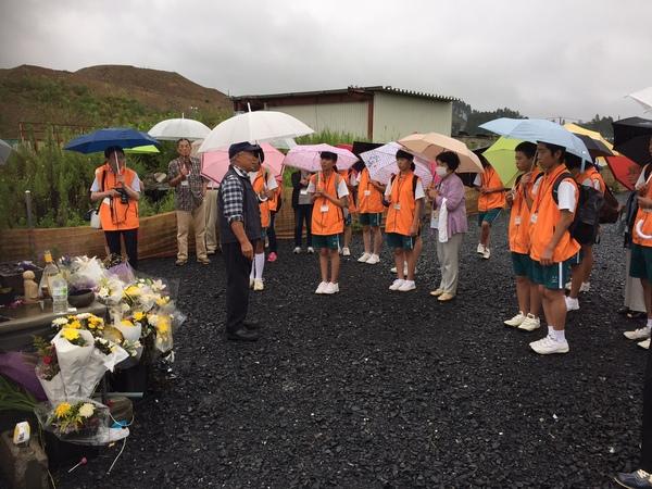 雨の中、傘をさしてジュニアボランティアの生徒が紺色ののベストを着た男性の周りに集まって話を聞いており、生徒達の前には沢山の献花が供えられている様子の写真
