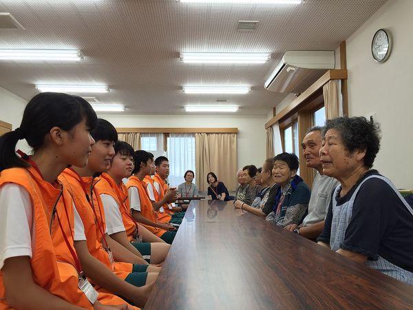 ジュニアボランティアの生徒が机を挟んで対面で住民の方が座っており、住民の年配の女性が話をしているのを聞いている様子の写真
