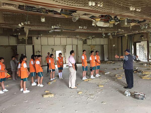 ジュニアリーダーの生徒達が、震災当日に芸能大会が開催された会場の視察を行っており、ベストを着た男性が両手を広げて説明をしており、建物の中は当時のままで、床はコンクリートで壁紙もはがれ、天井も破れてしまっている様子の写真