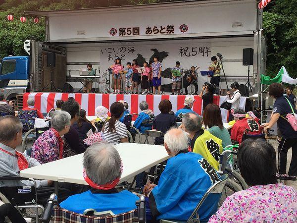 第5回あらと祭りが開催されており、舞台上にはギターやキーボートの演奏に合わせて5名の男の子と女の子が歌を歌っており、老人ホームの入所者が祭りの法被を着て見ている様子の写真