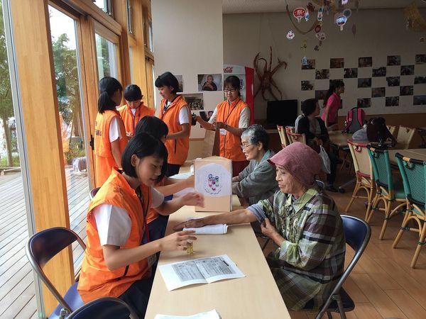 ジュニアボランティアの女子生徒が机に座って、年配の女性の手にアロマを付けてマッサージをしようとしている様子の写真