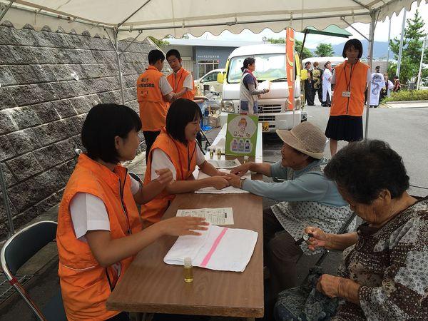 テントの下に「指マッサージ（アロマセラピー）」のブースを設けており、ジュニアボランティアの女子生徒が年配の女性の手をマッサージしている様子の写真