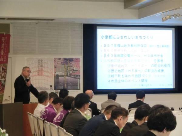 13名ほどの人が座っている前で大きな白いスクリーンに文字が映され、その横でマイクを持った男性が話をしている写真