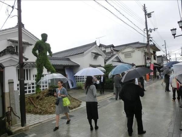 緑の人型のオブジェの前の道路を雨の中、傘をさして歩く10名ほどの男女の写真