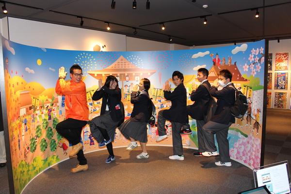 祭りの絵が書かれている壁面の前で学生6人が、手を叩きながら片足をあげてポーズをとっている写真