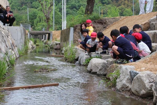 中央に川が流れていて、川沿いで子どもたちが手にコップを持ってしゃがみこんでいる写真