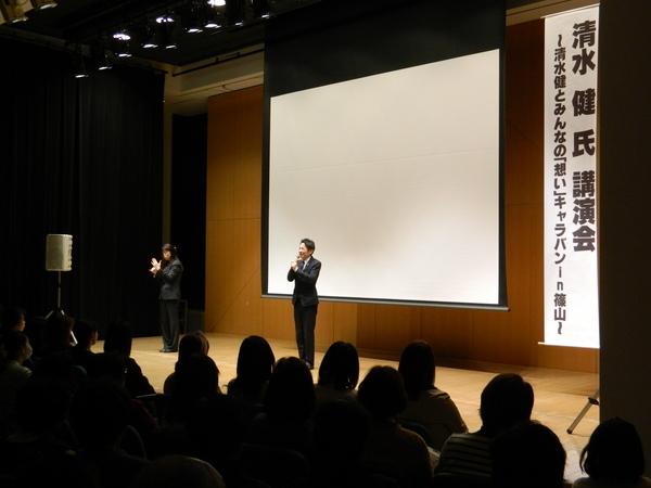 清水 健さんが壇上に上がってスクリーンの前でマイクを持って話をしており、左奥には手話通訳者が同時通訳をしている様子の写真