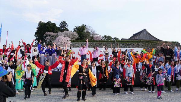 篠山城の屋根の見えるステージ上とその周りで、色とりどりの衣装を着た参加者が両手を上に挙げて歓声を上げている様子の写真