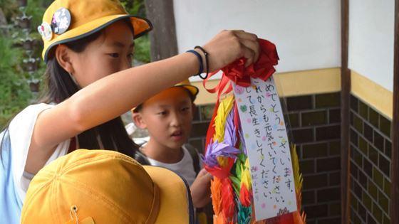 小学生が米寿祝に作ったメッセージ付きの千羽鶴を持っている写真