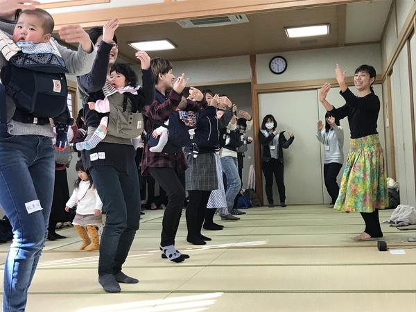 赤ちゃんを抱っこひもで抱っこしたお母さん達が横一列に並んで、講師の畠 ゆうこさんのフラダンスの振り付けを見ながら楽しそうに踊っている様子の写真