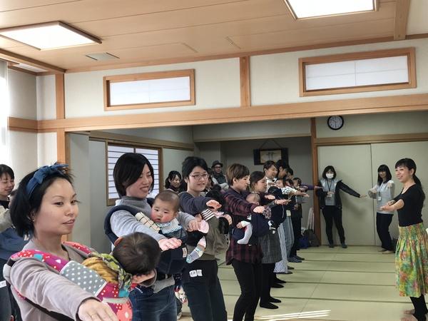 赤ちゃんを抱っこひもで前に抱っこして、講師の畠 ゆうこ先生の振り付けをまねて、両腕を肩の高さに右側に挙げて踊りを踊っている様子の写真