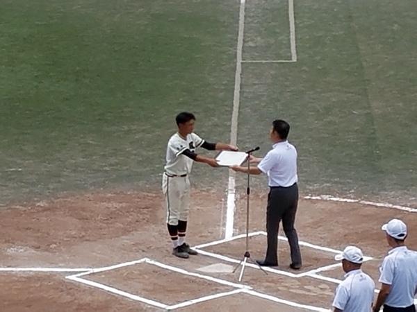 グランドのバッター席で、選手の一人と白いポロシャツの男性が額に入った賞状を手渡ししている様子の写真