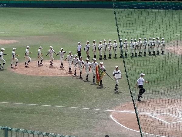 グランドに篠山鳳鳴高校軟式野球部の選手が一列に並んで行進しており、先頭の選手が額に入った賞状を持ち、次の選手が優勝旗を持って行進している様子の写真