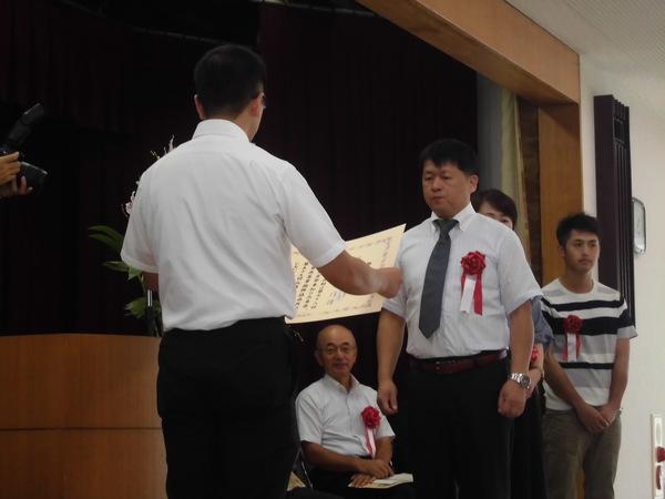 酒井 一行さんが胸に赤い胸章を付けており、賞状を読まれ表彰されている様子の写真