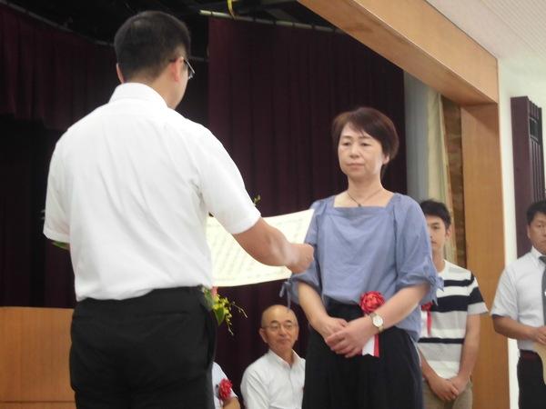 株式会社諏訪園の代表の女性が腰に赤い胸章を付けており、賞状を読まれ表彰されている様子の写真