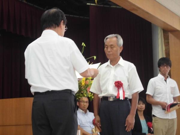 柳澤 廣明さんが胸に赤い胸章を付けており、賞状を読まれ表彰されている様子の写真