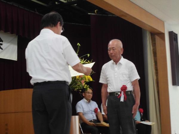 安井 雅彦さんが胸に赤い胸章を付けており、賞状を読まれ表彰されている様子の写真