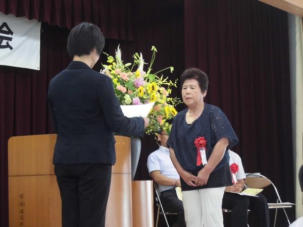 酒井 八重子さんが胸に赤い胸章を付けており、賞状を読まれ表彰されている様子の写真