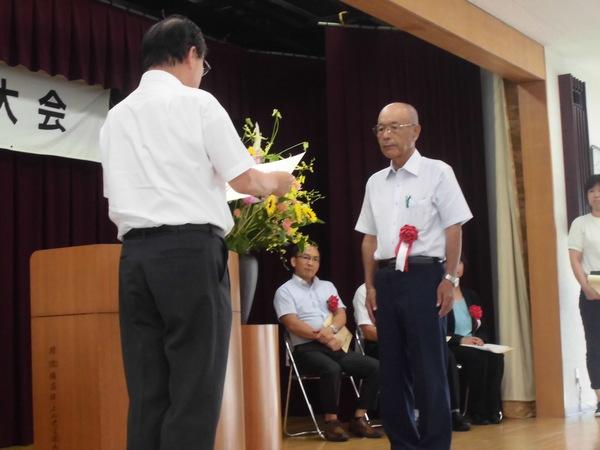 丹波篠山茶生産組合の代表の男性が胸に赤い胸章を付けており、賞状を読まれ表彰されている様子の写真