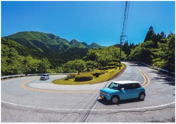 道路のカーブを曲がっている水色と灰色の車があり、遠くには青々とした木々が写っている写真