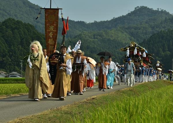 沿道の両脇に稲が栄え、獅子舞などの被り物を持った人々の後方に駕籠を持った駕籠舁が歩いている写真