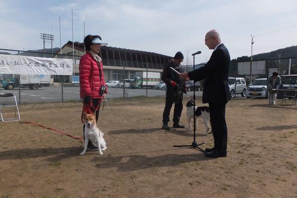 赤いダウンジャケットを着た女性と雑種の犬に市長が認定書を読んでいる写真