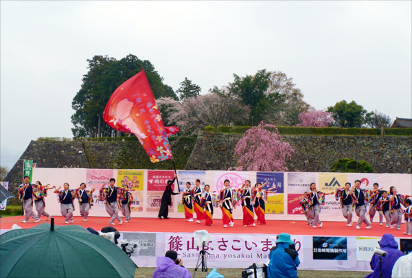傘をさす人、合羽を着る人達がが見守る中ステージで踊っている人達の写真