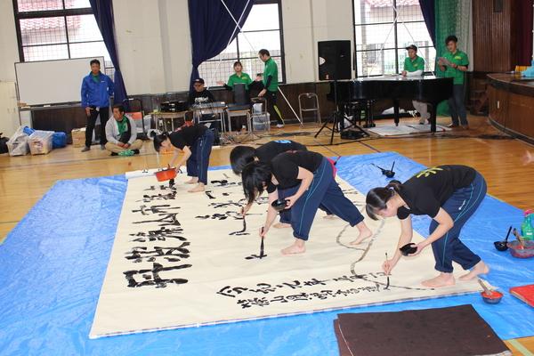 4人の女子学生が床に置かれた大きな紙に書道パフォーマンス行っている様子の写真