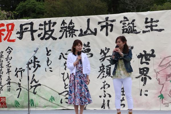 祝丹波篠山誕生と書道パフォーマンスで書かれた作品の前でマイクを持ち話をしている女性2人の写真