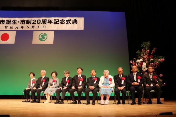 舞台上で受賞者の皆様が左胸に赤い花のリボンを付けて、椅子に座っている写真