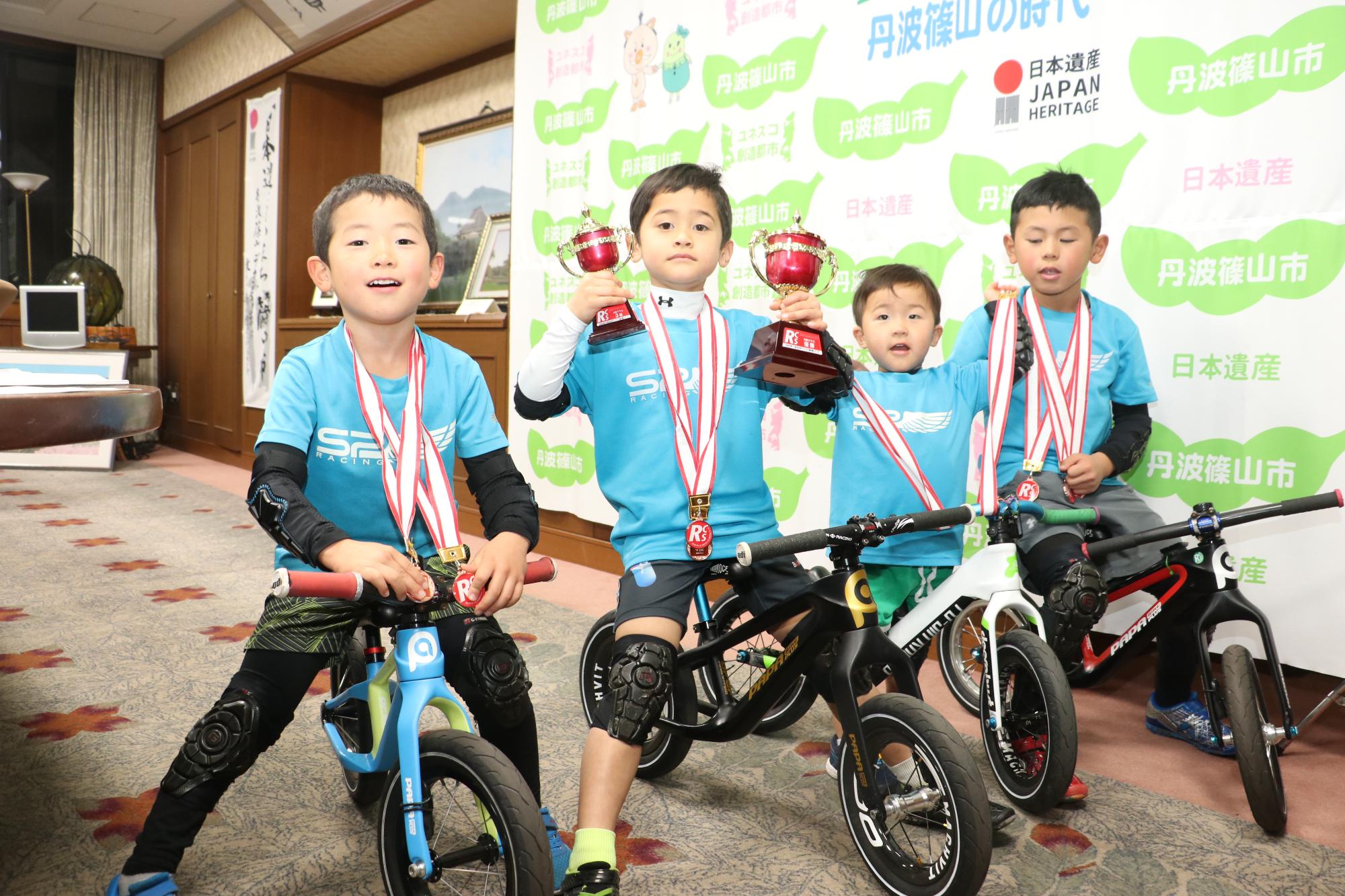 4人の幼児が首にメダルをかけて自転車に乗りトロフィーを掲げている