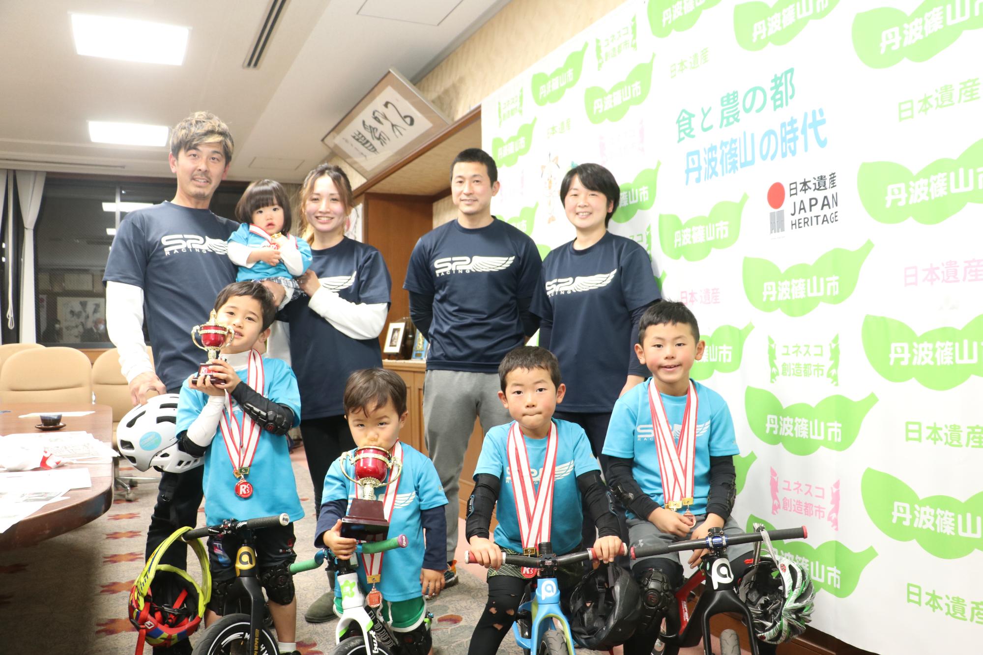 首にメダルをかけて自転車にまたがる4人の男児とその両親4人が並んでいる
