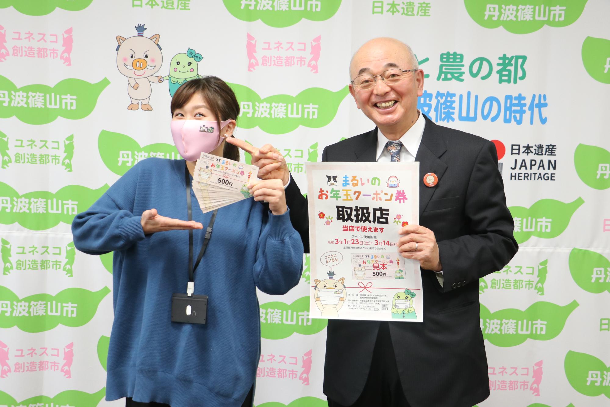 市長が笑顔でチラシを持ち、隣で女性職員がクーポン券を紹介している