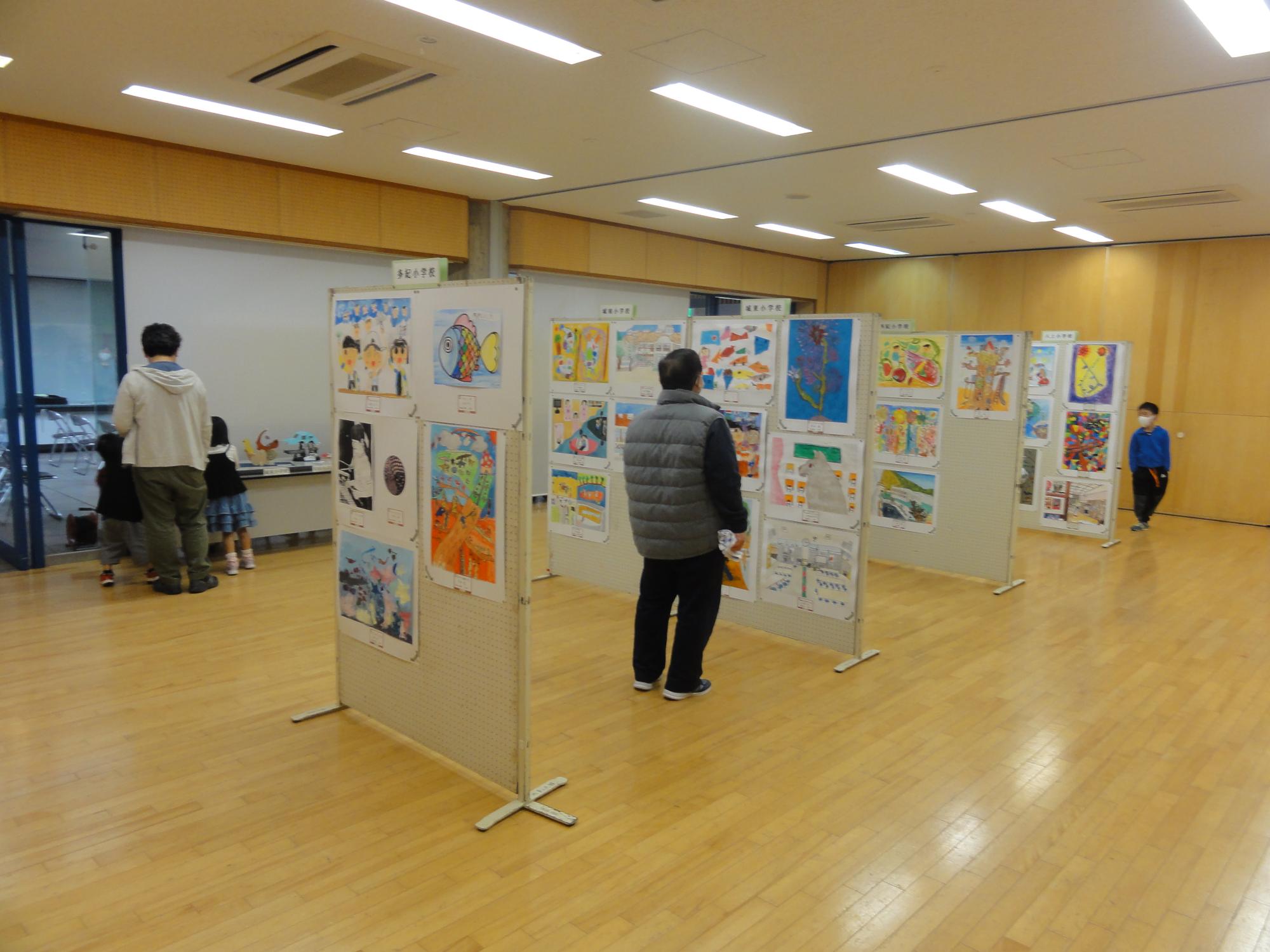 パネル4枚に小学生の絵画、机には工作を展示していて、来場者が作品を眺めている。