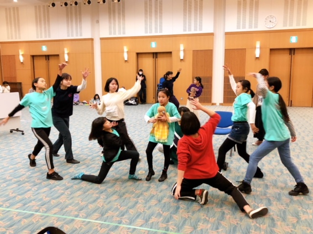 片方の手をあげもう片方の手を下げて複数の女の子が人形を持った一人の女の子の周りに集まっている写真